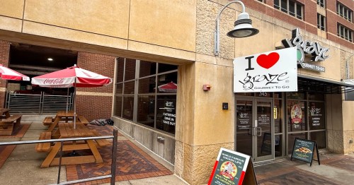 The exterior patio of Graze Gourmet To-Go featuring wooden picnic tables with red umbrellas and a large sign with a heart graphic near the entrance.