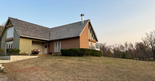 A large house with tan siding and brick sections sitting on a dry grassy hill under a clear sky.