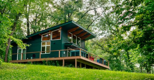 A modern dark green cabin with large windows and a wooden deck, nestled on a grassy hill surrounded by lush green trees.