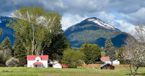 Quaint, brightly colored homes sit before looming mountains draped in imposing clouds. The surrounding nature is green with a few trees behind the buildings.