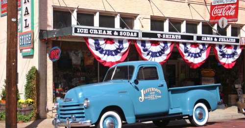 A vintage blue pickup truck with whitewall tires is parked in front of a historic general store decorated with red, white, and blue bunting, with signs advertising ice cream, sodas, T-shirts, and jams and jellies.