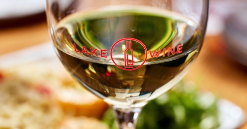 a wine glass with the logo for lake wine is filled with white wine is set in the foreground, and a plate with greens and other food is on a white plate in the background