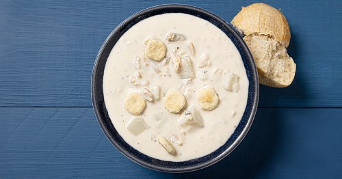 a bowl of New England clam chowder topped with oyster crackers sits on a navy blue wood table next to a rill broken in half