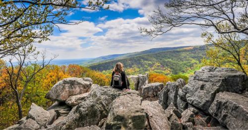 A hiker perched on a rocky cliff overlooking the Appalachian Mountains, glowing with autumn leaves on a partly cloudy day.