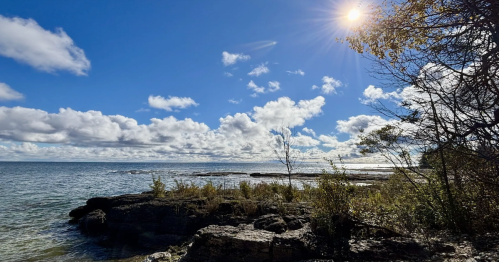 Bright sunshine bursts over the shimmering waters of Lake Michigan, illuminating the rocky coast and scattered clouds at Lynd Point on a breezy day.