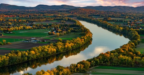 A calm, wide river cuts in between green farmland dotted with small buildings at dusk. The river is lined with trees changing for fall and mountains loom large in the distance.