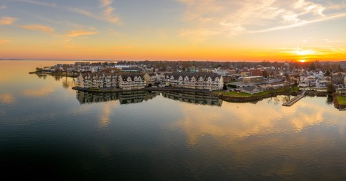 An orange sunset glows over the outline of a coastal town with residences sitting right next to the water. The water remains still to a point of near perfect reflection.