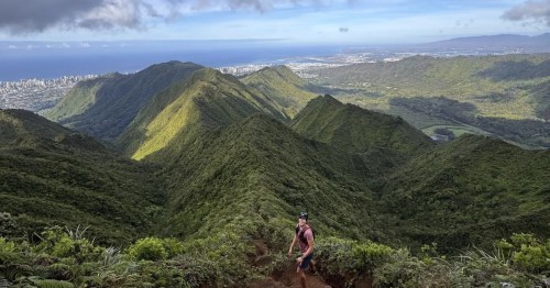 Hiker standing on a narrow ridgeline trail with sweeping views of sharp green mountain peaks and the ocean in the distance.