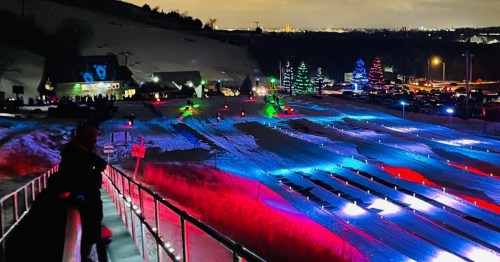 Night tubing hill illuminated with vibrant neon green, pink, and blue lights overlooking a city.