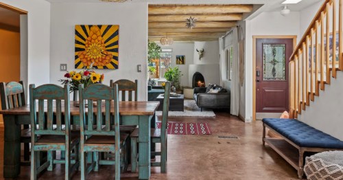 a dining table surrounded by turquoise-colored wood chairs and beyond that is a living room with a fire lit in the kiva fireplace