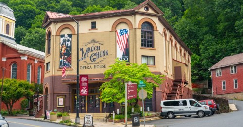 An old fashioned tan and brick theater sits in front of a vibrantly green forest next to other crisp, brick buildings.