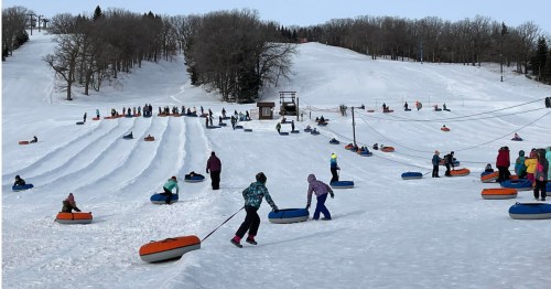 Many people pulling blue and orange snow tubes up a multi-lane snowy tubing hill lined with trees.