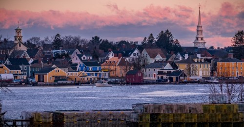 Dawn colors the sky pastel oranges, pinks, and purples as a town with colonial and shingle style architecture begins to wake up. A boat chugs along a calm river.