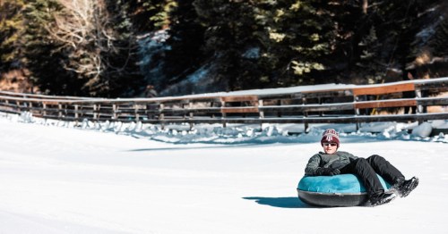 A child sitting upright in a blue snow tube, gliding across a snowy tubing lane with a wooden fence and evergreen forest in the background.