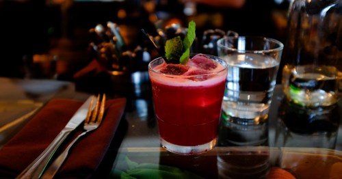 view of the Beet Down, a cocktail at Root Down at Denver International Airport, sitting on a bar next ton a glass of water, cloth napkin, and fork and knife