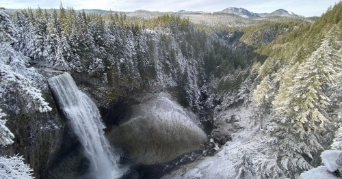 Wide view of a tall waterfall cascading into a forested gorge, with snow-covered pine trees lining the canyon and soft winter light illuminating the landscape.