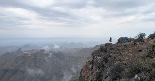 Two hikers standing near the edge of a high desert overlook with layered mountain ranges fading into the distance beneath overcast skies.