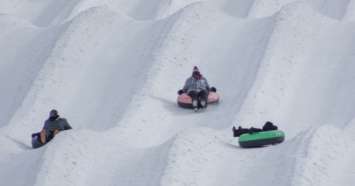 Three people on inflatable tubes are positioned at different points while sliding down snowy hill lanes.