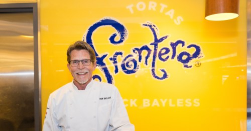 Celebrity chef Rick Bayless stands in front of a yellow wall with the name of his restaurant, tortas frontera on it