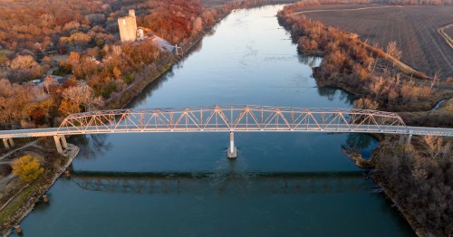 An aerial view of the Brownville Bridge spanning the Missouri River, illuminated by golden hour sunlight with autumn trees and a grain elevator visible on the riverbank.