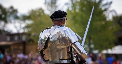A knight in shiny silver plate armor seen from behind, holding a sword before a blurred crowd.