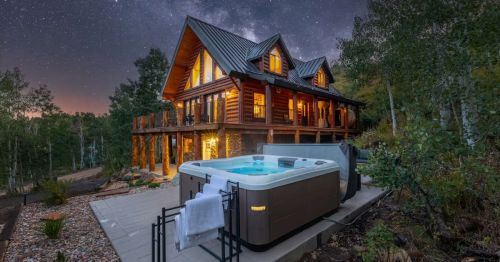 A log cabin in Utah, tucked on a hill with a luxurious hot tub, surrounded by bare towering trees underneath the Milky Way galaxy.