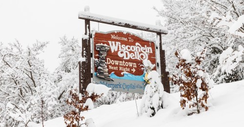 The wooden "Welcome to Wisconsin Dells Downtown" sign is covered in fresh snow and surrounded by frost-covered trees, highlighting the city's title as "The Waterpark Capital of the World."