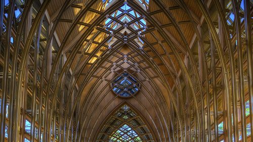 A modern wooden chapel with tall arches, large windows, and rows of benches, illuminated by natural light.