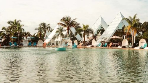 A serene pool scene with palm trees, lounge chairs, and modern pyramid-shaped buildings in the background.