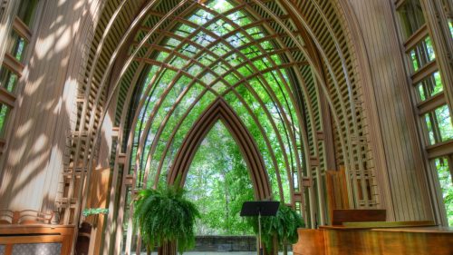 A serene wooden chapel with arched ceilings, surrounded by greenery and featuring a piano in the foreground.