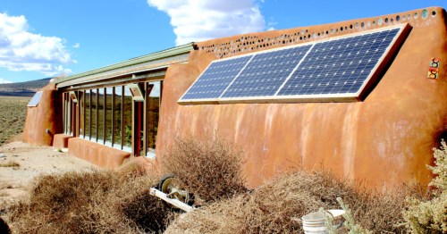 Exterior of Encounter Earthship in El Prado, NM, with adobe walls, solar panels, and a glass front under a blue sky.