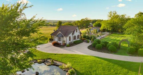 Exterior of English cottage Airbnb in Harrison, Arkansas, with grass and gardens in the foreground and trees in the background on a sunny day.