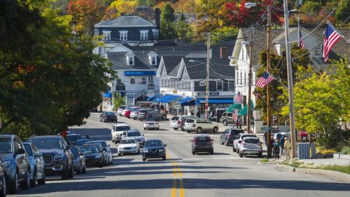 A scenic street lined with cars, featuring shops and colorful autumn trees in the background. American flags are visible.