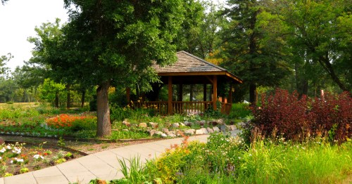 A wooden gazebo surrounded by colorful flower beds and lush green trees at the International Peace Garden.