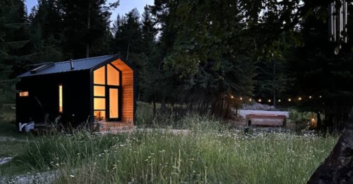 A Japandi-inspired tiny house with yellow light coming from its massive windows, and nestled in a forest setting.