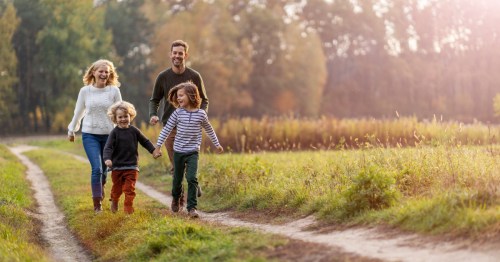 A young family smiles and laughs as they run down a dirt road in a green field. Tall, green trees are visible behind them.