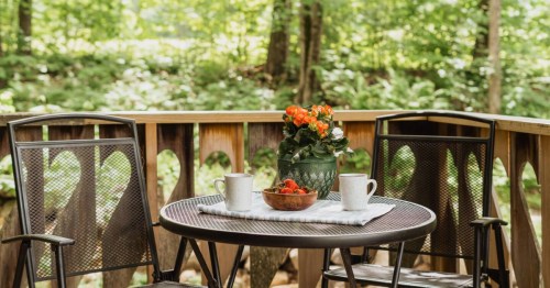 Two black cast iron chairs and a table sit on the wrap-around deck of The Chalet Swiss-style AIrbnb property in Nelson, New Hampshire. Flowers, coffee, and a bowl of strawberries are placed on the table.