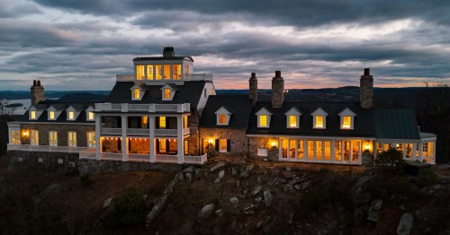 A large English-accented mansion with a stone exterior on a hill, illuminated by lights inside at sunset.