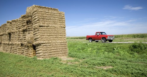Red pickup truck drives past massive hay bale stacks along the rural Old Lincoln Highway in Nebraska.