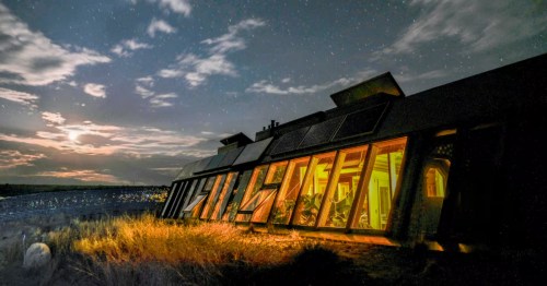 Spectacular Earthship at night with glowing interior lights, solar panels, and a star-filled sky in Taos County.