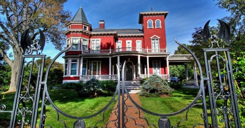 A dark, red Victorian mansion featuring a wrought iron fence decorated with bats and spiders.