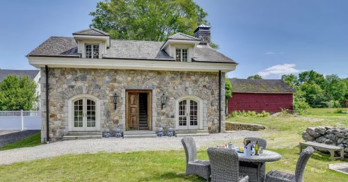 A sunny exterior view of the Stone House Airbnb château in Stonington, Connecticut. A wicker table and four chairs sit on the lawn in front of the stone home.