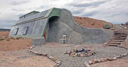Exterior of the Taos Mesa Studio Earthship with solar panels, a glass facade, and adobe walls under a cloudy New Mexico sky.