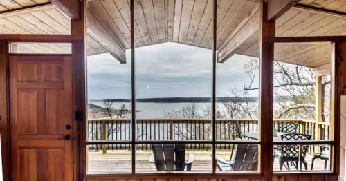 Large floor-to-ceiling windows inside a wooden cabin overlooking a calm lake and a wrap-around deck.
