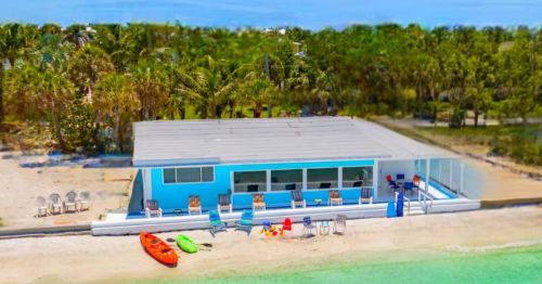 A blue Airbnb vacation rental in Florida on the water has a sandy beach out back and various chairs and colorful water sports equipment lined up.