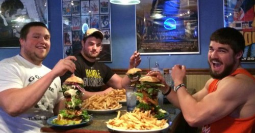 Three men at a restaurant table posing with towering burgers and large plates of french fries.