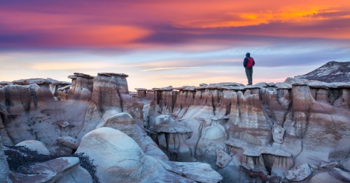 A lone hiker with a red backpack stands atop otherworldly hoodoos at Bisti/De-Na-Zin Wilderness near Farmington, New Mexico, beneath a dramatic pastel sunset sky streaked with pink, purple, and orange.