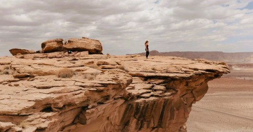 Person standing on the edge of a rocky cliff with a vast desert landscape along the cross-state trail to Skylight Arch in AZ.