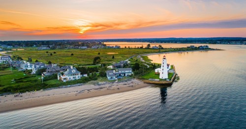 The sun sets on a flat stretch of land where white houses reside on a patch of green grass bordering a calm ocean overseen by a white lighthouse.