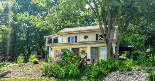 Charming two-story cottage with bright yellow doors, surrounded by lush greenery and flowers.
